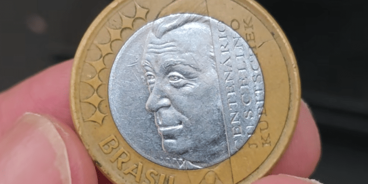 Close detalhado da moeda de 1 real JK sendo segurada por dedos. A face prateada exibe o perfil de Juscelino Kubitschek com a inscrição "CENTENÁRIO JUSCELINO KUBITSCHEK", enquanto o anel dourado externo mostra a palavra "BRASIL" e estrelas decorativas.