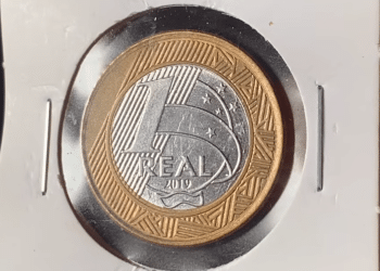 Close de uma moeda de 1 real do ano de 2019 protegida em um suporte de papel branco. A imagem foca no lado do valor, exibindo o número 1 grande, a palavra REAL e o ano 2019 logo abaixo, com detalhes da constelação do Cruzeiro do Sul ao fundo.