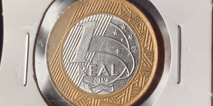 Close de uma moeda de 1 real do ano de 2019 protegida em um suporte de papel branco. A imagem foca no lado do valor, exibindo o número 1 grande, a palavra REAL e o ano 2019 logo abaixo, com detalhes da constelação do Cruzeiro do Sul ao fundo.