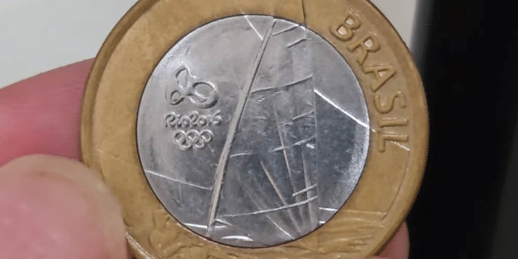 Close-up de uma moeda de 1 real das Olimpíadas Rio 2016 (tema vela), sendo segurada por dedos, mostrando a borda dourada e o centro prateado com o desenho do barco.