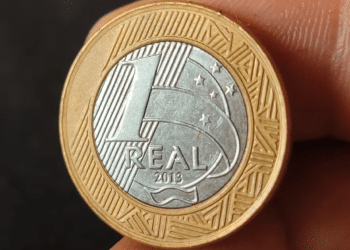 Close de uma moeda de 1 real do ano de 2013 sendo segurada pelos dedos, mostrando detalhadamente o valor de face e os elementos de segurança no reverso prateado.