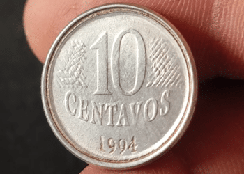 Close-up de uma moeda de 10 centavos de real do ano de 1994, feita de aço inox, sendo segurada pelos dedos de uma pessoa. A imagem foca no reverso, onde o número "10" e a palavra "CENTAVOS" estão centralizados, com a data "1994" logo abaixo. É possível notar detalhes da cunhagem e pequenos sinais de circulação.