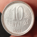 Close-up de uma moeda de 10 centavos de real do ano de 1994, feita de aço inox, sendo segurada pelos dedos de uma pessoa. A imagem foca no reverso, onde o número "10" e a palavra "CENTAVOS" estão centralizados, com a data "1994" logo abaixo. É possível notar detalhes da cunhagem e pequenos sinais de circulação.