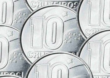 Close de várias moedas prateadas de 10 Cruzeiros sobrepostas em um plano detalhado. É possível ver claramente o número 10 e a palavra "CRUZEIROS" gravados em metal brilhante com estrelas ao fundo.