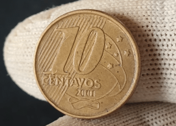 Close-up de uma mão usando luva de tecido branca segurando uma moeda de 10 centavos de real do ano de 2001. A imagem foca no valor de face "10 centavos" e na data, destacando os detalhes de conservação e os traços da cunhagem.
