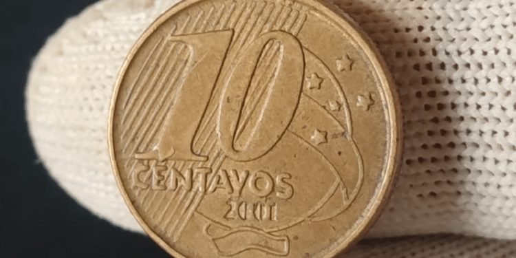 Close-up de uma mão usando luva de tecido branca segurando uma moeda de 10 centavos de real do ano de 2001. A imagem foca no valor de face "10 centavos" e na data, destacando os detalhes de conservação e os traços da cunhagem.