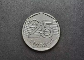 Vista em close-up da face anversa de uma moeda de 25 centavos de real, mostrando o valor numérico "25" e a palavra "CENTAVOS" em relevo, em fundo escuro e liso.