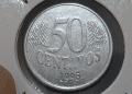 Close-up de uma moeda de 50 centavos de real do ano 1995 em um suporte de proteção branco. A face exibida mostra o valor "50", a palavra "CENTAVOS" abaixo e o ano "1995" na base, com detalhes de linhas diagonais ao fundo.