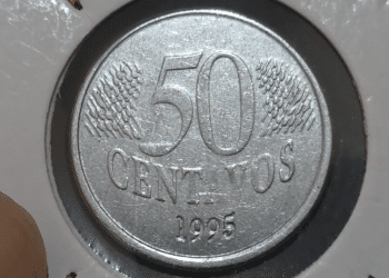 Close-up de uma moeda de 50 centavos de real do ano 1995 em um suporte de proteção branco. A face exibida mostra o valor "50", a palavra "CENTAVOS" abaixo e o ano "1995" na base, com detalhes de linhas diagonais ao fundo.
