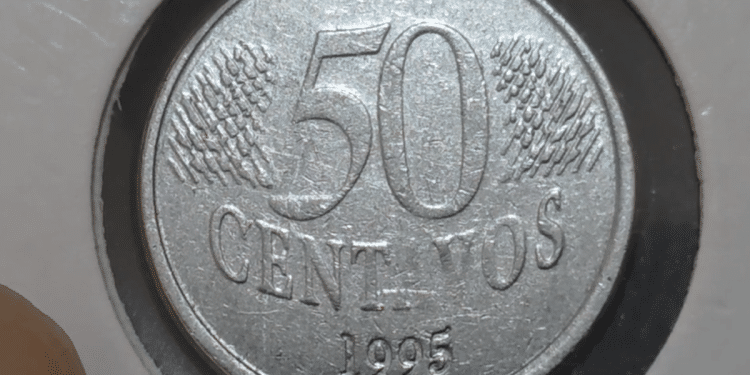 Close-up de uma moeda de 50 centavos de real do ano 1995 em um suporte de proteção branco. A face exibida mostra o valor "50", a palavra "CENTAVOS" abaixo e o ano "1995" na base, com detalhes de linhas diagonais ao fundo.