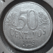 Close-up de uma moeda de 50 centavos de real do ano 1995 em um suporte de proteção branco. A face exibida mostra o valor "50", a palavra "CENTAVOS" abaixo e o ano "1995" na base, com detalhes de linhas diagonais ao fundo.