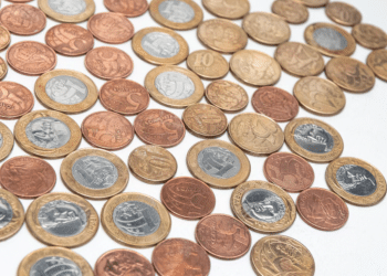 Diversas moedas de Real brasileiro de diferentes valores (1 Real, 50 centavos, 10 centavos) espalhadas sobre uma superfície branca lisa. As moedas apresentam diferentes estados de oxidação e brilho, com destaque para as bordas serrilhadas e o relevo do cunho.