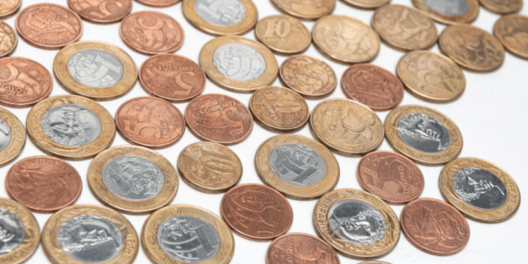 Diversas moedas de Real brasileiro de diferentes valores (1 Real, 50 centavos, 10 centavos) espalhadas sobre uma superfície branca lisa. As moedas apresentam diferentes estados de oxidação e brilho, com destaque para as bordas serrilhadas e o relevo do cunho.