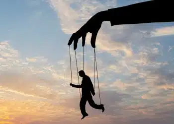 Silhueta de mão gigante controlando homem com fios contra céu ao entardecer