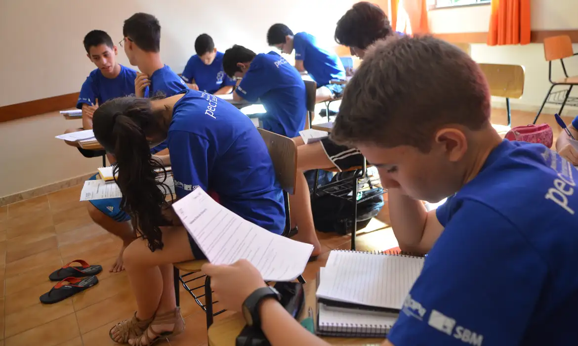 Estudantes sentados em sala de aula, vestindo uniformes azuis, concentrados na leitura e resolução de cadernos de prova da OBMEP.