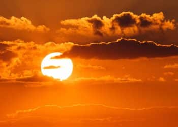Paisagem de um pôr do sol intenso com o céu em tons de laranja vibrante e nuvens escuras, simbolizando o aquecimento global e as ondas de calor extremo.
