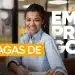 Jovem mulher negra sorridente com cabelos cacheados presos, vestindo camisa social azul claro, sentada em um escritório. Sobre a imagem, há o texto em destaque "VAGAS DE EMPREGO" em cores branco e laranja.
