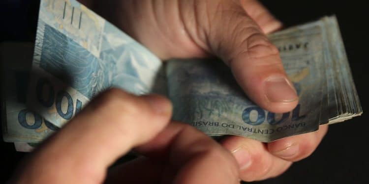 Beneficiário contando cédulas de R$ 100 representando antecipação do 13º salário do INSS que injeta R$ 78 bilhões na economia