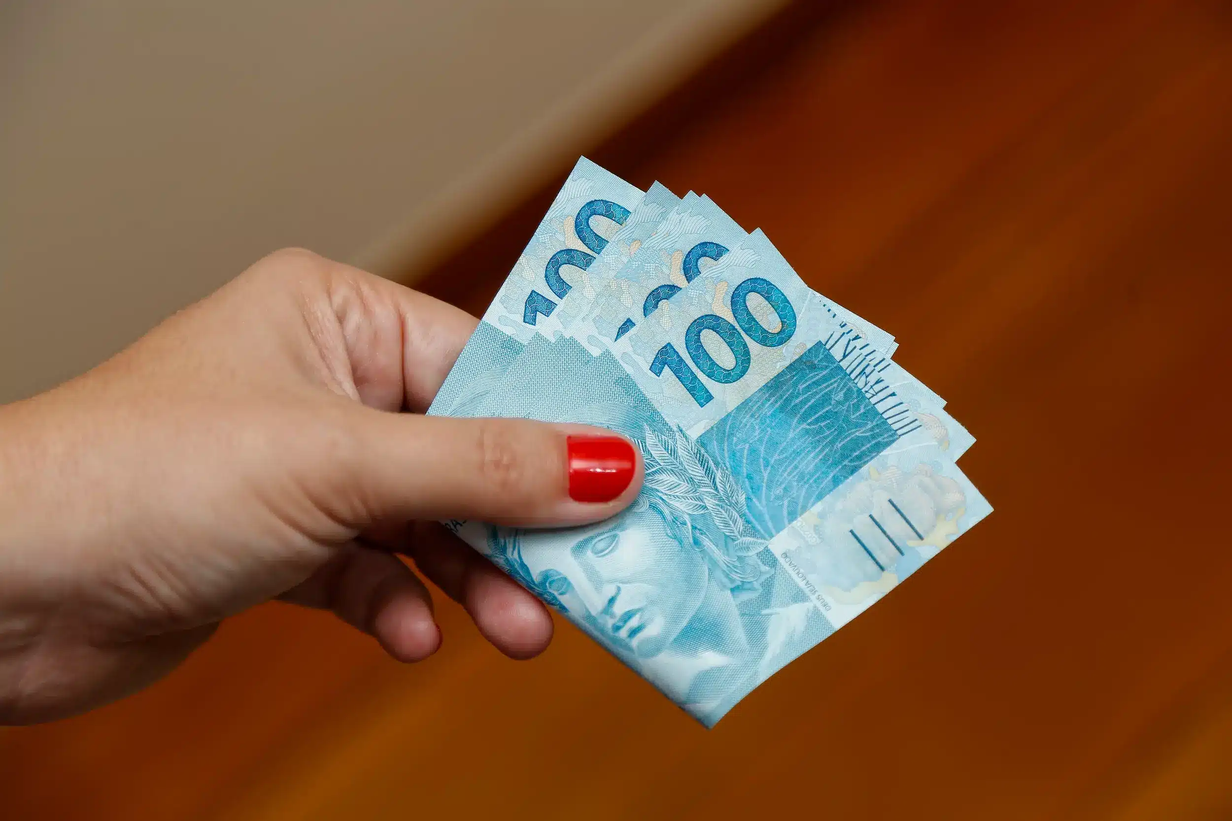 Pessoa segurando notas de cem reais representando o pagamento de R$ 2.200 do benefício financeiro.