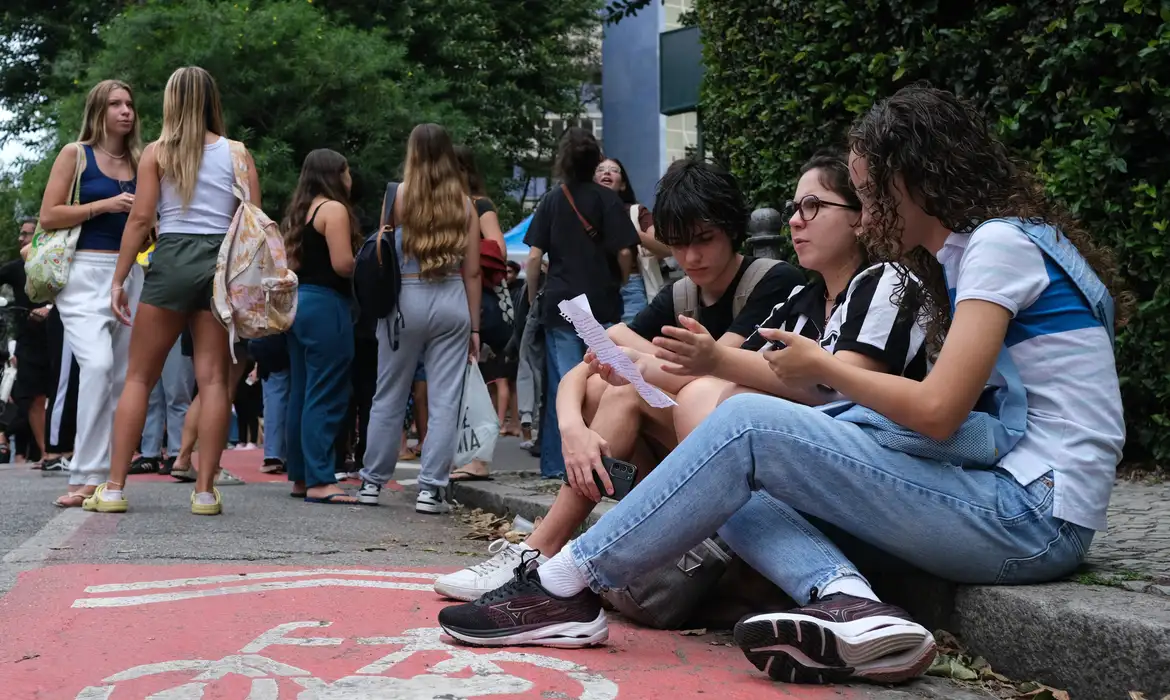 Jovens reunidos em calçada, alguns sentados lendo papéis, esperam início de prova em ambiente urbano