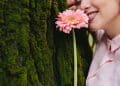 Mulher encostada em tronco de árvore com musgo segurando uma gérbera rosa próxima ao rosto, ilustrando a relação entre flores e personalidade