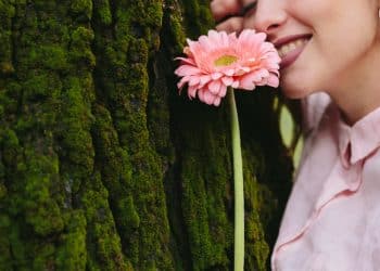 Mulher encostada em tronco de árvore com musgo segurando uma gérbera rosa próxima ao rosto, ilustrando a relação entre flores e personalidade
