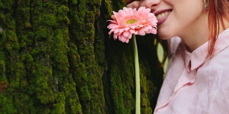 Mulher encostada em tronco de árvore com musgo segurando uma gérbera rosa próxima ao rosto, ilustrando a relação entre flores e personalidade
