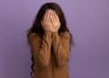 Uma mulher de cabelos longos e castanhos usando uma blusa marrom, cobrindo o rosto com as duas mãos em um gesto de timidez ou vergonha, contra um fundo roxo liso.