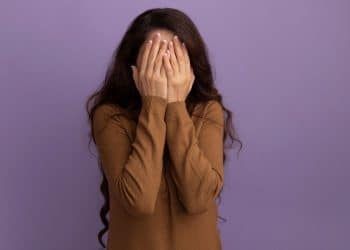 Uma mulher de cabelos longos e castanhos usando uma blusa marrom, cobrindo o rosto com as duas mãos em um gesto de timidez ou vergonha, contra um fundo roxo liso.