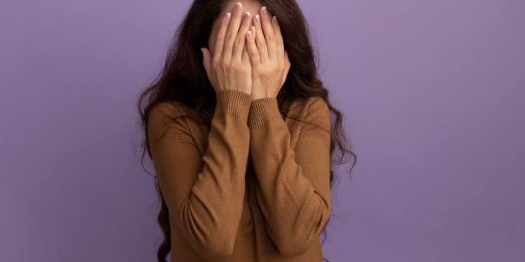 Uma mulher de cabelos longos e castanhos usando uma blusa marrom, cobrindo o rosto com as duas mãos em um gesto de timidez ou vergonha, contra um fundo roxo liso.