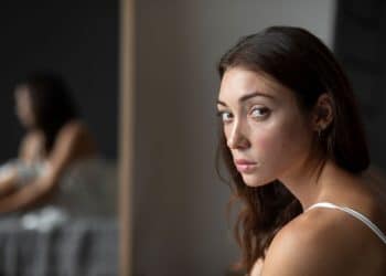 Retrato em close de uma mulher jovem de cabelos castanhos ondulados e olhar firme e direto, transmitindo compostura e serenidade. Ao fundo, seu reflexo em um espelho aparece levemente desfocado, simbolizando a introspecção e o domínio próprio. Ela veste uma regata branca simples em um ambiente de tons neutros.