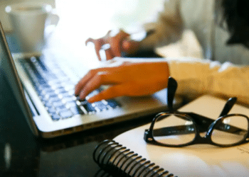 Pessoa digitando em notebook com óculos e caderno na mesa representando estudo online