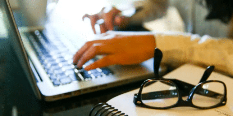 Pessoa digitando em notebook com óculos e caderno na mesa representando estudo online