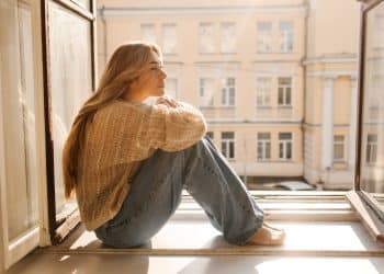 Mulher com cabelo loiro, vestindo suéter e jeans, sentada na janela em um dia ensolarado, refletindo sobre a solitude e o tempo sozinha.
