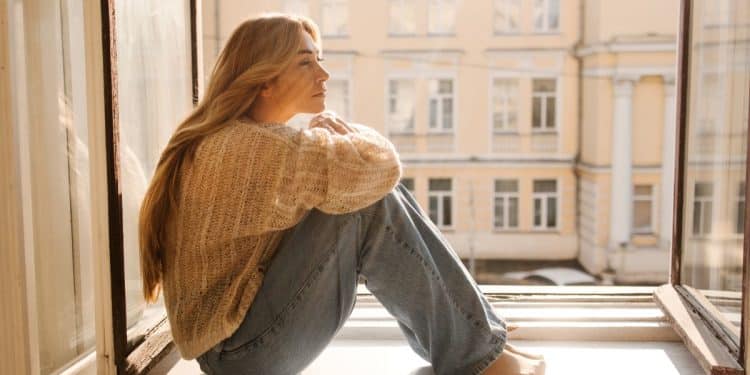 Mulher com cabelo loiro, vestindo suéter e jeans, sentada na janela em um dia ensolarado, refletindo sobre a solitude e o tempo sozinha.