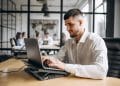 Homem jovem de barba e camisa social branca trabalhando concentrado em um notebook em um escritório moderno. Ao fundo, colegas de trabalho conversam em um ambiente corporativo com divisórias de vidro.
