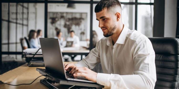 Homem jovem de barba e camisa social branca trabalhando concentrado em um notebook em um escritório moderno. Ao fundo, colegas de trabalho conversam em um ambiente corporativo com divisórias de vidro.
