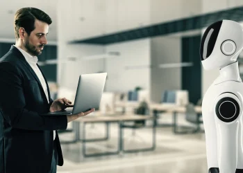 Profissional utilizando notebook diante de robô humanoide em escritório moderno, representando a colaboração entre inteligência artificial e habilidades humanas no mercado de trabalho.