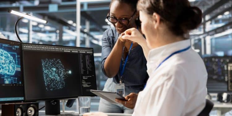 Duas mulheres em um escritório moderno de tecnologia celebram com um toque de mãos ("fist bump"). Uma delas, negra e com óculos, segura um tablet, enquanto a outra está sentada à frente de dois monitores que exibem gráficos de redes neurais e um cérebro digital.