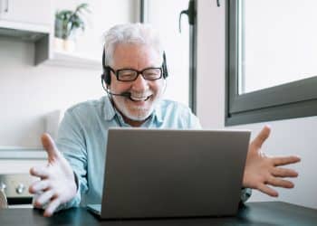 Homem grisalho sorrindo com headset em frente a notebook durante chamada de vídeo