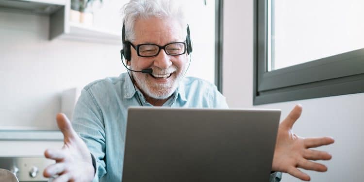 Homem grisalho sorrindo com headset em frente a notebook durante chamada de vídeo