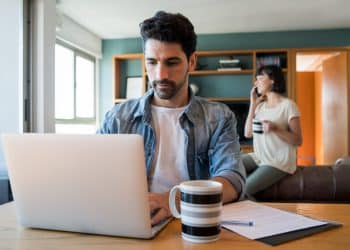 Homem trabalhando de casa no notebook com caneca de café na mesa da sala