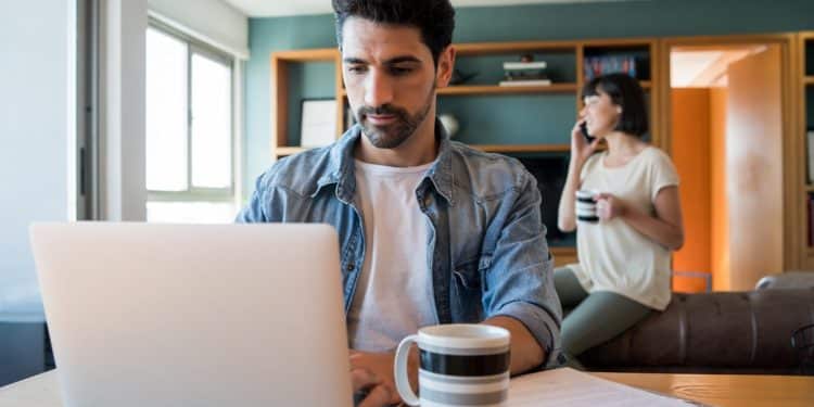 Homem trabalhando de casa no notebook com caneca de café na mesa da sala