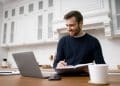 Homem sorrindo trabalhando em home office com laptop fone de ouvido e caderno representando profissões remotas bem remuneradas