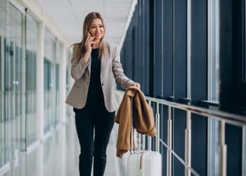 Profissional elegante falando ao celular em corredor de aeroporto com mala de viagem e casaco no braço representando carreiras que permitem viajar