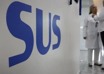 Letreiro azul SUS em parede, com médicos de branco conversando ao fundo em hospital