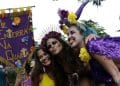 Três mulheres sorridentes em um bloco de Carnaval de rua segurando um estandarte roxo com a frase "ME ENTERRA NA QUARTA". A mulher à esquerda veste amarelo, a do centro usa uma tiara de flores roxas e a da direita tem cabelos azuis e capa de lantejoulas roxas.