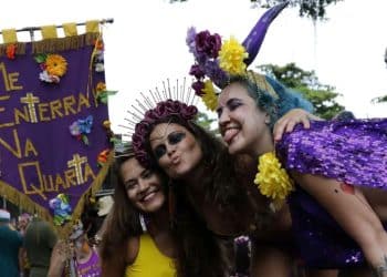 Três mulheres sorridentes em um bloco de Carnaval de rua segurando um estandarte roxo com a frase "ME ENTERRA NA QUARTA". A mulher à esquerda veste amarelo, a do centro usa uma tiara de flores roxas e a da direita tem cabelos azuis e capa de lantejoulas roxas.