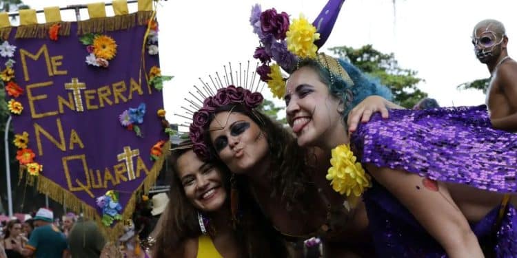 Três mulheres sorridentes em um bloco de Carnaval de rua segurando um estandarte roxo com a frase "ME ENTERRA NA QUARTA". A mulher à esquerda veste amarelo, a do centro usa uma tiara de flores roxas e a da direita tem cabelos azuis e capa de lantejoulas roxas.