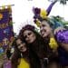 Três mulheres sorridentes em um bloco de Carnaval de rua segurando um estandarte roxo com a frase "ME ENTERRA NA QUARTA". A mulher à esquerda veste amarelo, a do centro usa uma tiara de flores roxas e a da direita tem cabelos azuis e capa de lantejoulas roxas.